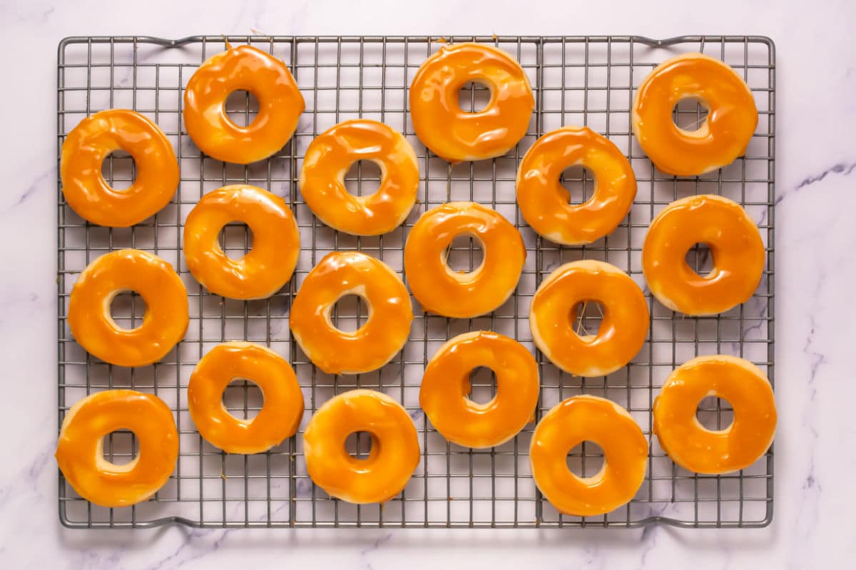 A cooling rack holds 20 round donuts coated with shiny orange glaze, arranged neatly in rows on a white marble surface, reminiscent of the look of Somoa Copy Cat Cookies.