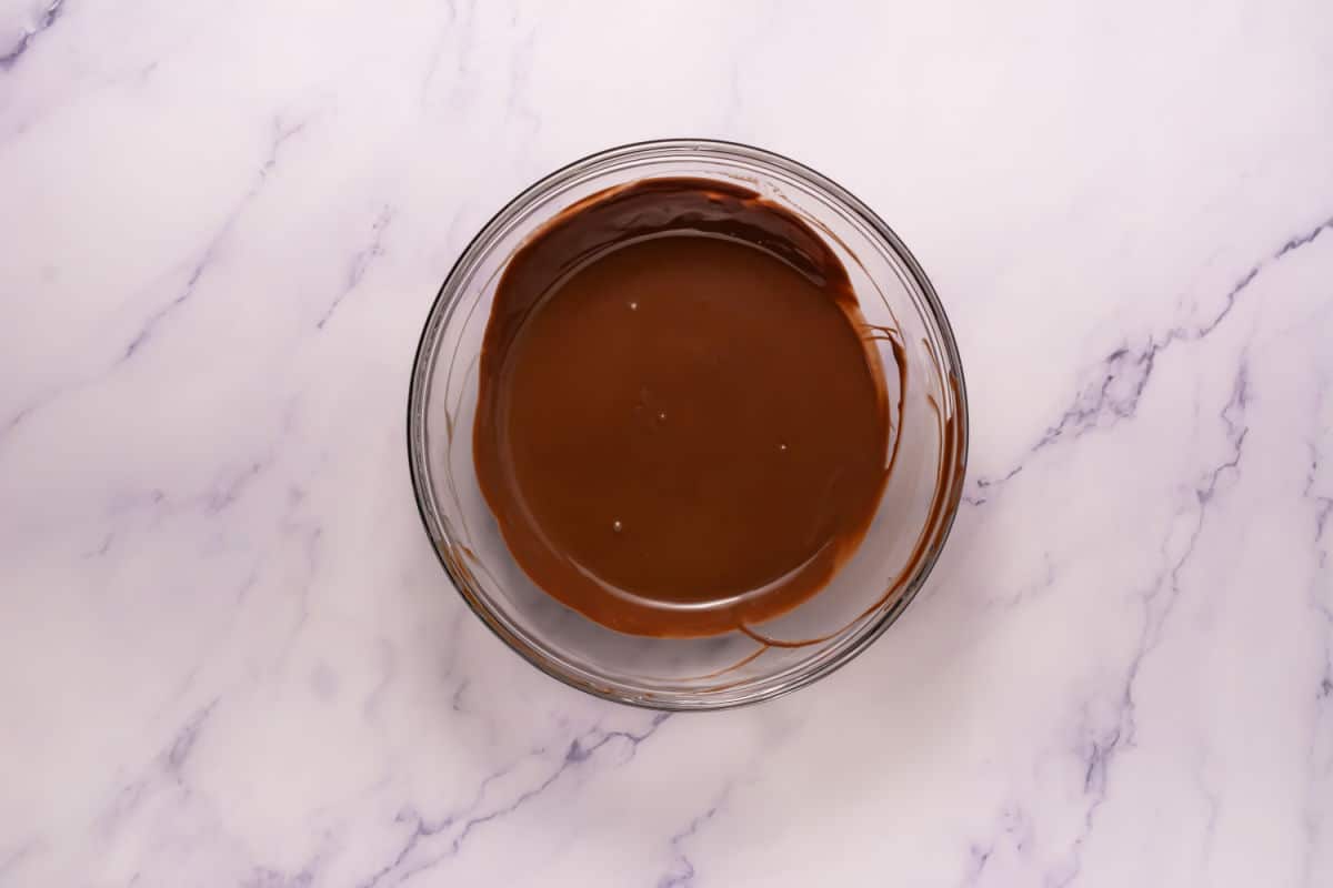 A glass bowl filled with smooth, melted chocolate sits on a white marble surface, ready to coat Somoa Copy Cat Cookies.