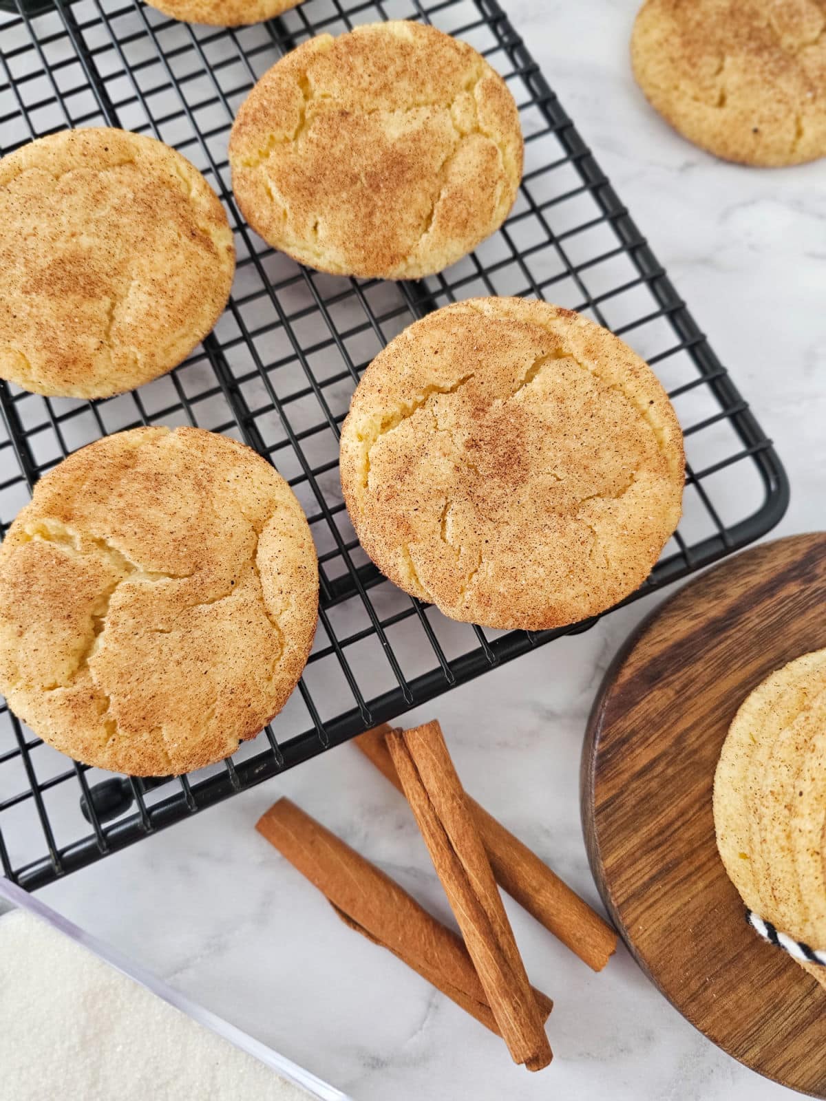 Freshly baked snickerdoodle cookies cool on a black rack, with two cinnamon sticks and an extra snickerdoodle cookie on a wooden board, all arranged atop a white marble surface.