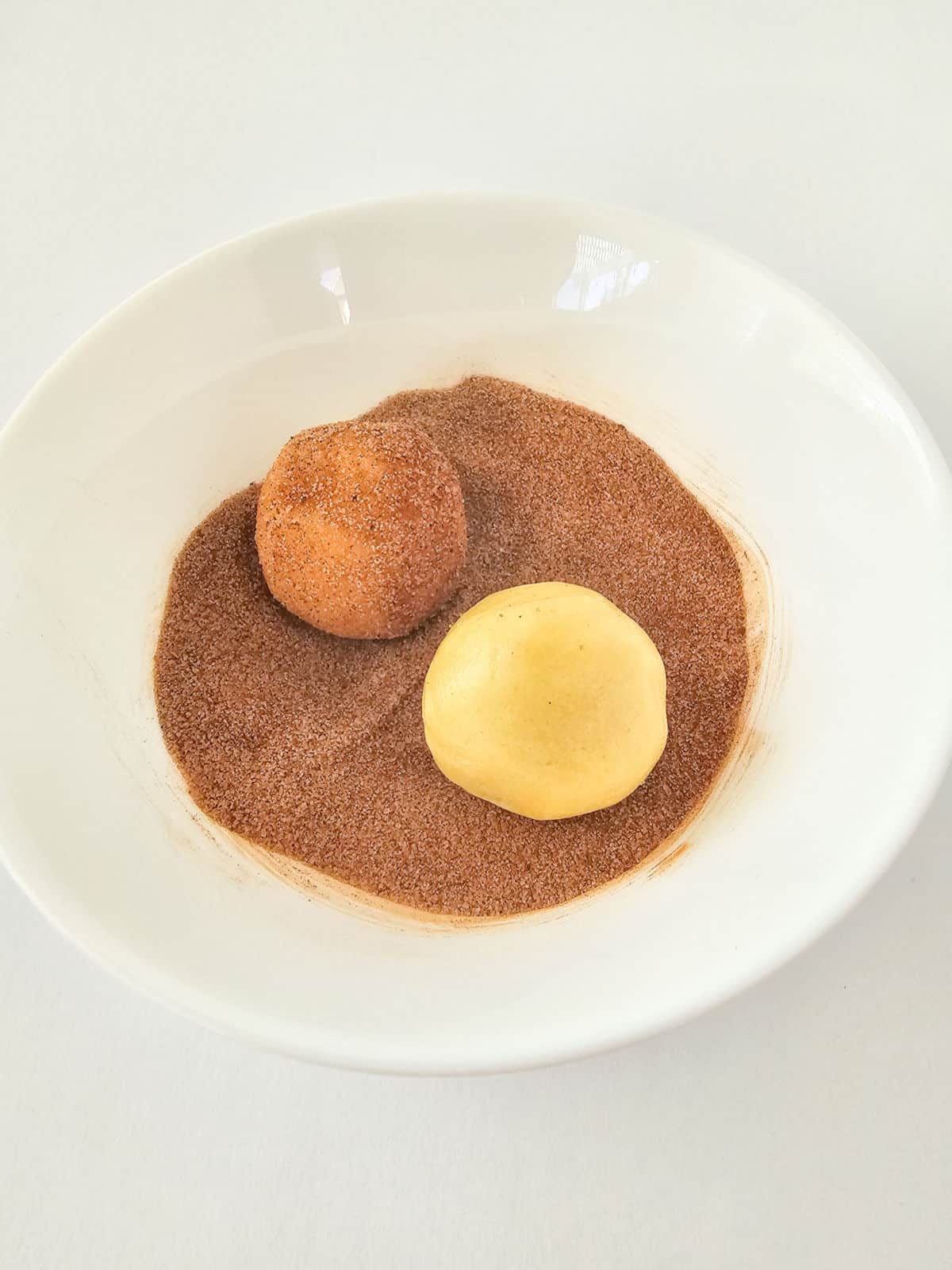 A white bowl containing a ball of snickerdoodle cookie dough being coated in cinnamon sugar, next to another uncoated ball of dough, on a white background.
