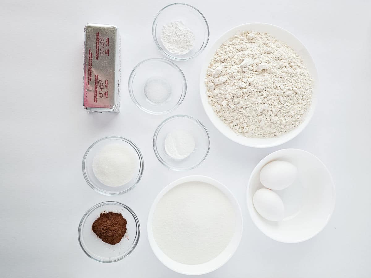 Top-down view of baking ingredients on a white surface, perfect for making snickerdoodle cookies: flour, eggs, butter, cocoa powder, sugar, and small bowls of baking powder, baking soda, and salt.