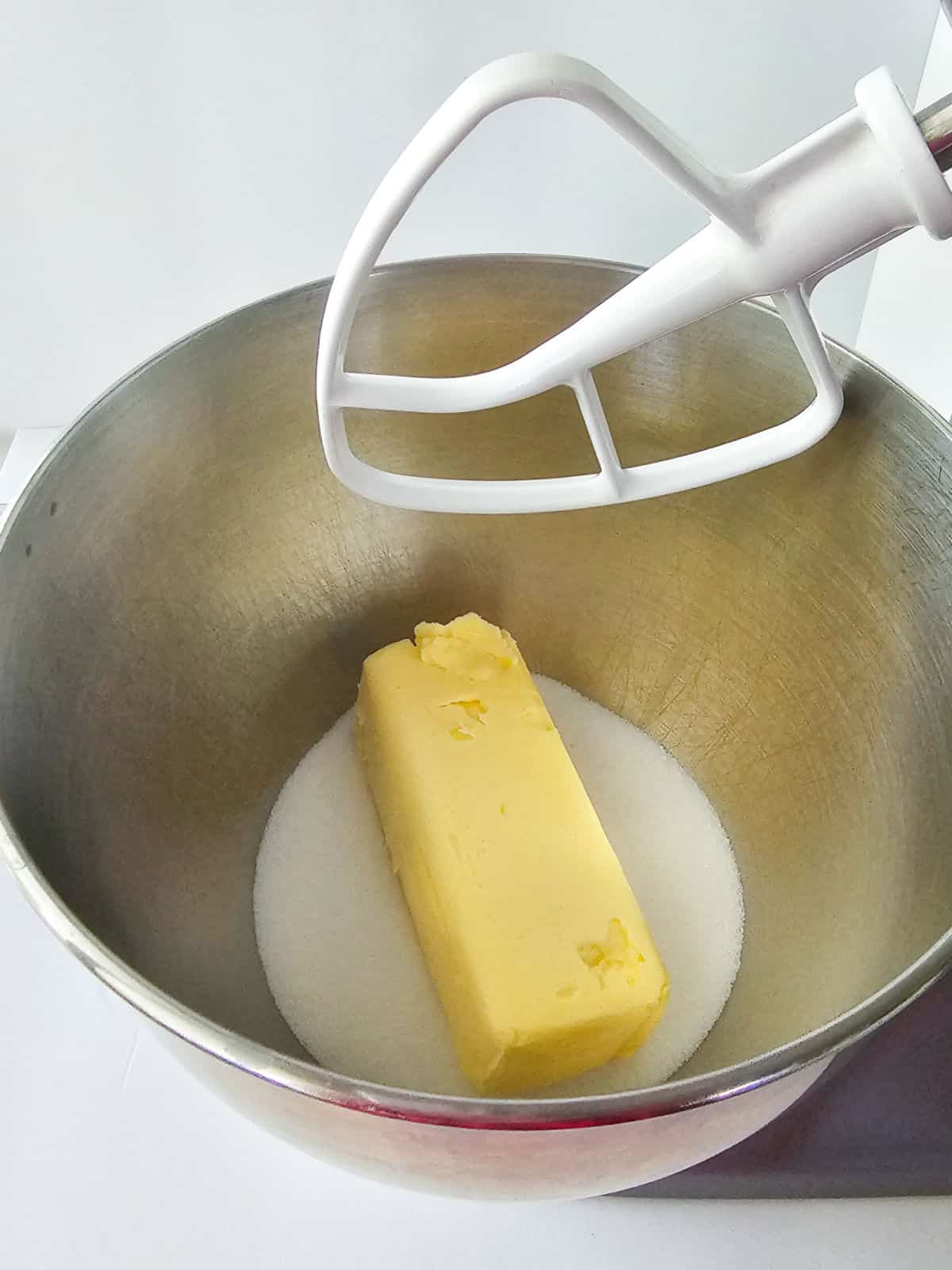 A stick of butter and granulated sugar sit in a metal mixing bowl, with a white stand mixer paddle attachment above—ingredients ready to be mixed for delicious snickerdoodle cookies.