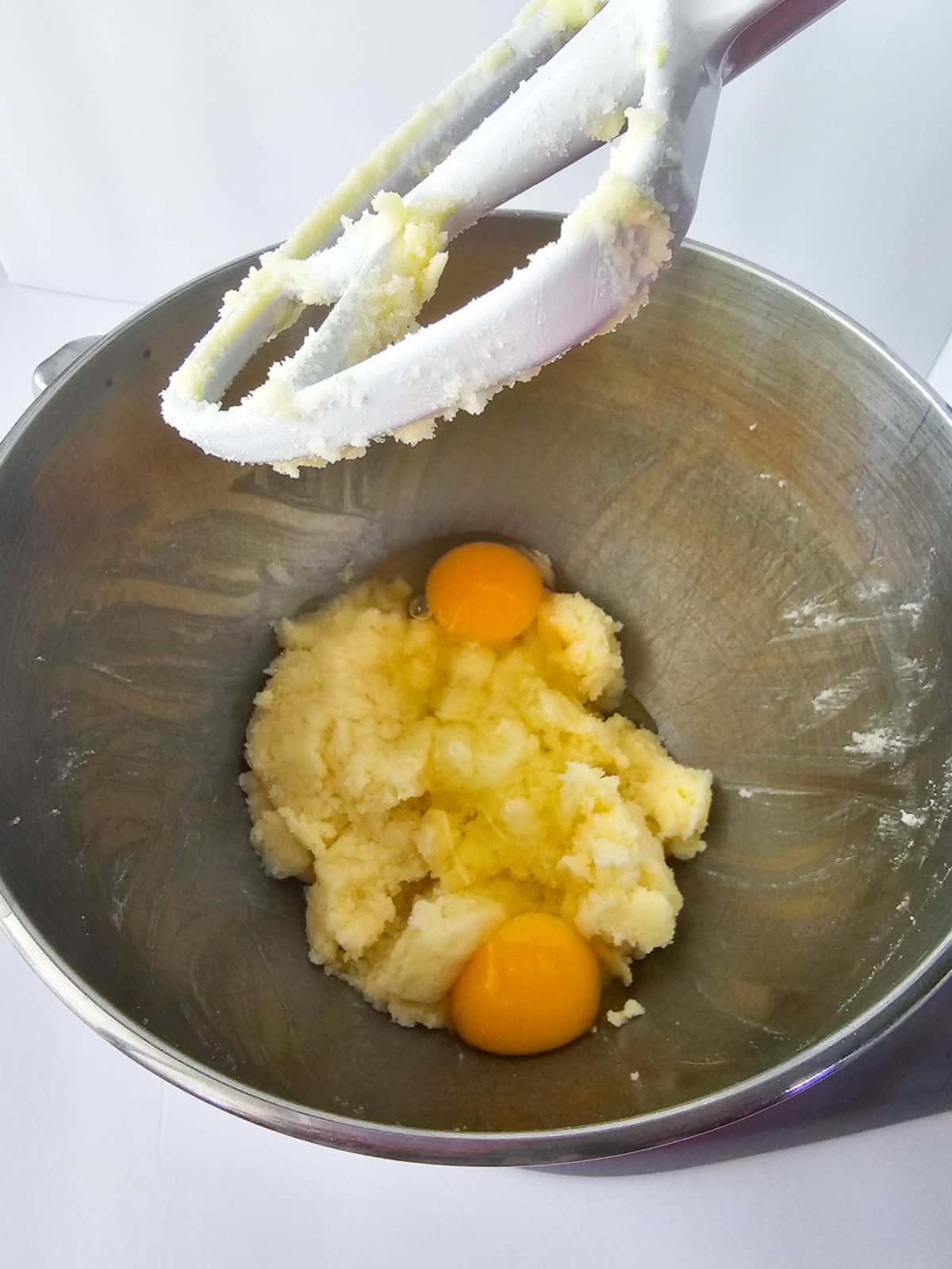 A metal mixing bowl contains creamed butter and sugar with two raw eggs on top—ingredients ready to become snickerdoodle cookies. An electric mixer paddle, coated with batter, is positioned above the bowl.