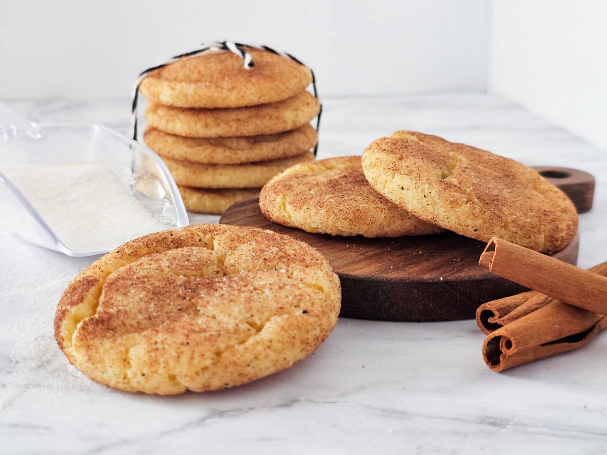 A stack of snickerdoodle cookies tied with string sits behind more snickerdoodle cookies on a wooden board. A scoop of sugar and cinnamon sticks are arranged nearby on a white marble surface.