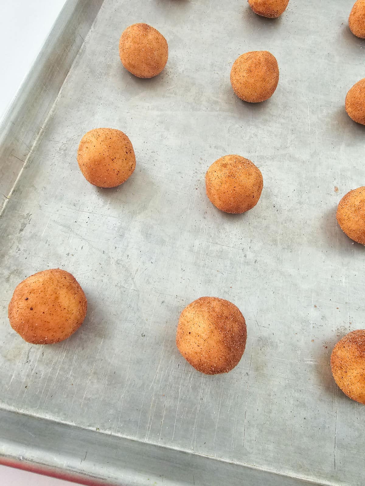 Round balls of snickerdoodle cookie dough coated in cinnamon sugar are spaced out on a metal baking sheet, ready to be baked.