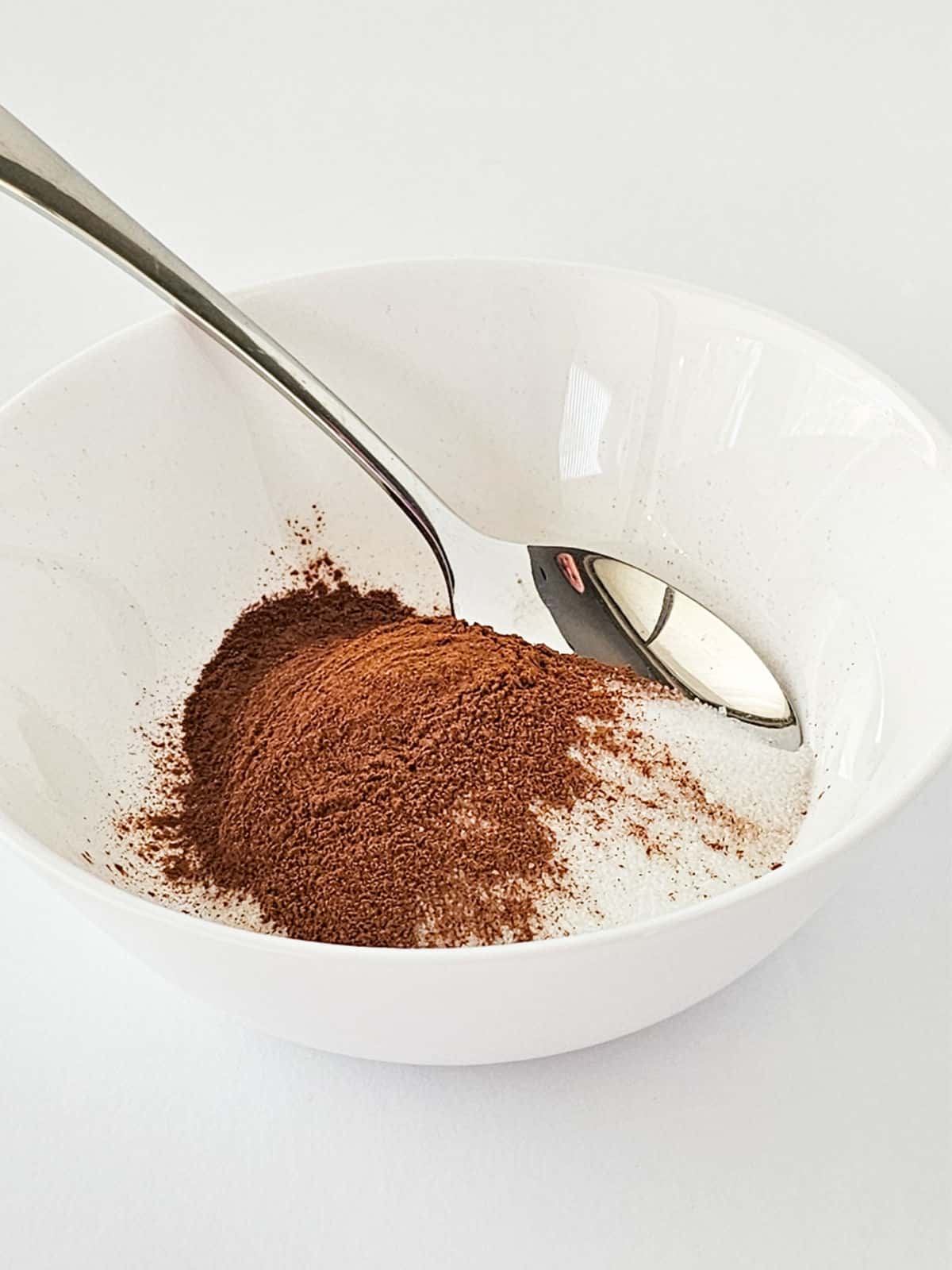 A white bowl containing a pile of brown cinnamon powder on top of white granulated sugar, with a metal spoon resting inside the bowl—perfect for making classic snickerdoodle cookies.