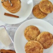 Two white plates with snickerdoodle cookies on a marble surface, one plate stacked and tied with string, the other with cookies spread out. Nearby are a cinnamon stick and a container of granulated sugar, highlighting the classic snickerdoodle flavor.