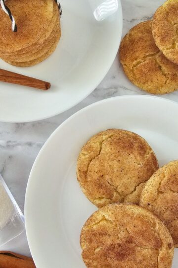 Two white plates with snickerdoodle cookies on a marble surface, one plate stacked and tied with string, the other with cookies spread out. Nearby are a cinnamon stick and a container of granulated sugar, highlighting the classic snickerdoodle flavor.