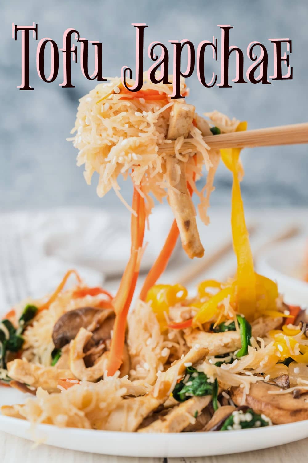 A plate of Tofu Japchae with Vermicelli, featuring stir-fried vegetables and noodles, is lifted by chopsticks. Carrots, mushrooms, and spinach are visible. The text “Tofu Japchae” appears at the top of the image.