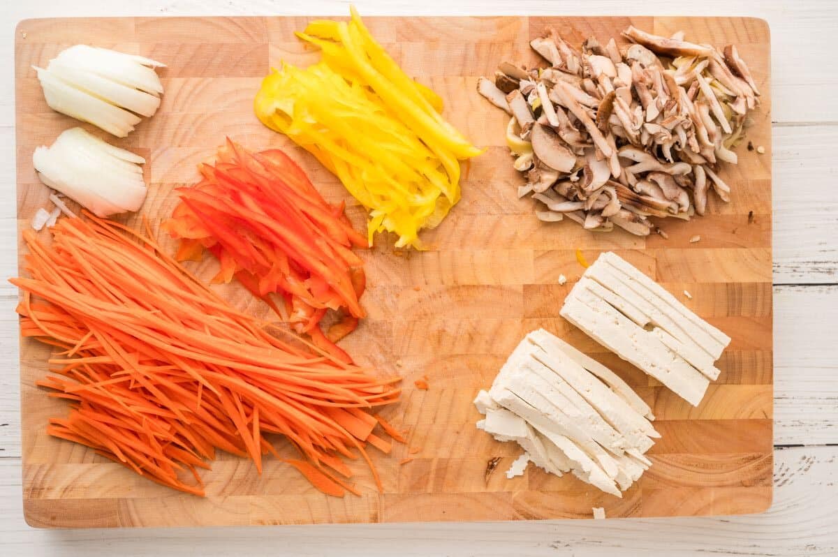A wooden cutting board with neatly arranged piles of sliced onion, yellow bell pepper, red bell pepper, carrots, mushrooms, and tofu—perfect ingredients for preparing Tofu Japchae with Vermicelli on a white surface.