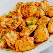 A white plate filled with golden-brown grilled chicken bites, coated in a glossy honey garlic sauce and garnished with chopped fresh herbs.