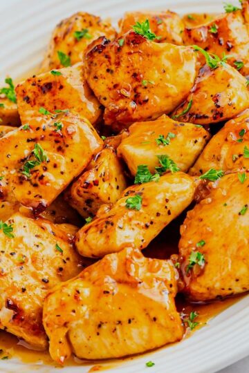 A white plate filled with golden-brown grilled chicken bites, coated in a glossy honey garlic sauce and garnished with chopped fresh herbs.