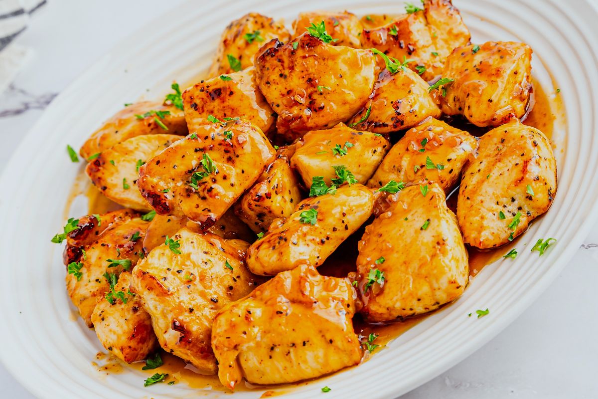 A white plate filled with golden-brown grilled chicken bites, coated in a glossy honey garlic sauce and garnished with chopped fresh herbs.