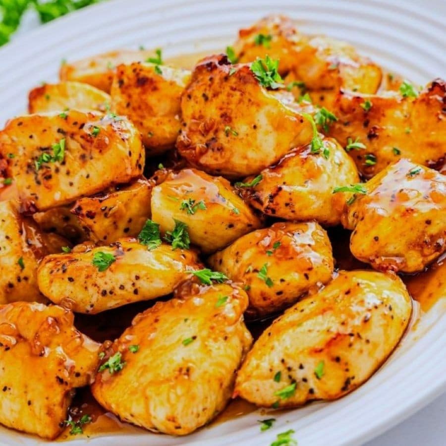 A plate of golden-brown, bite-sized chicken nuggets coated in a glossy honey garlic sauce, garnished with chopped parsley, served on a white dish.