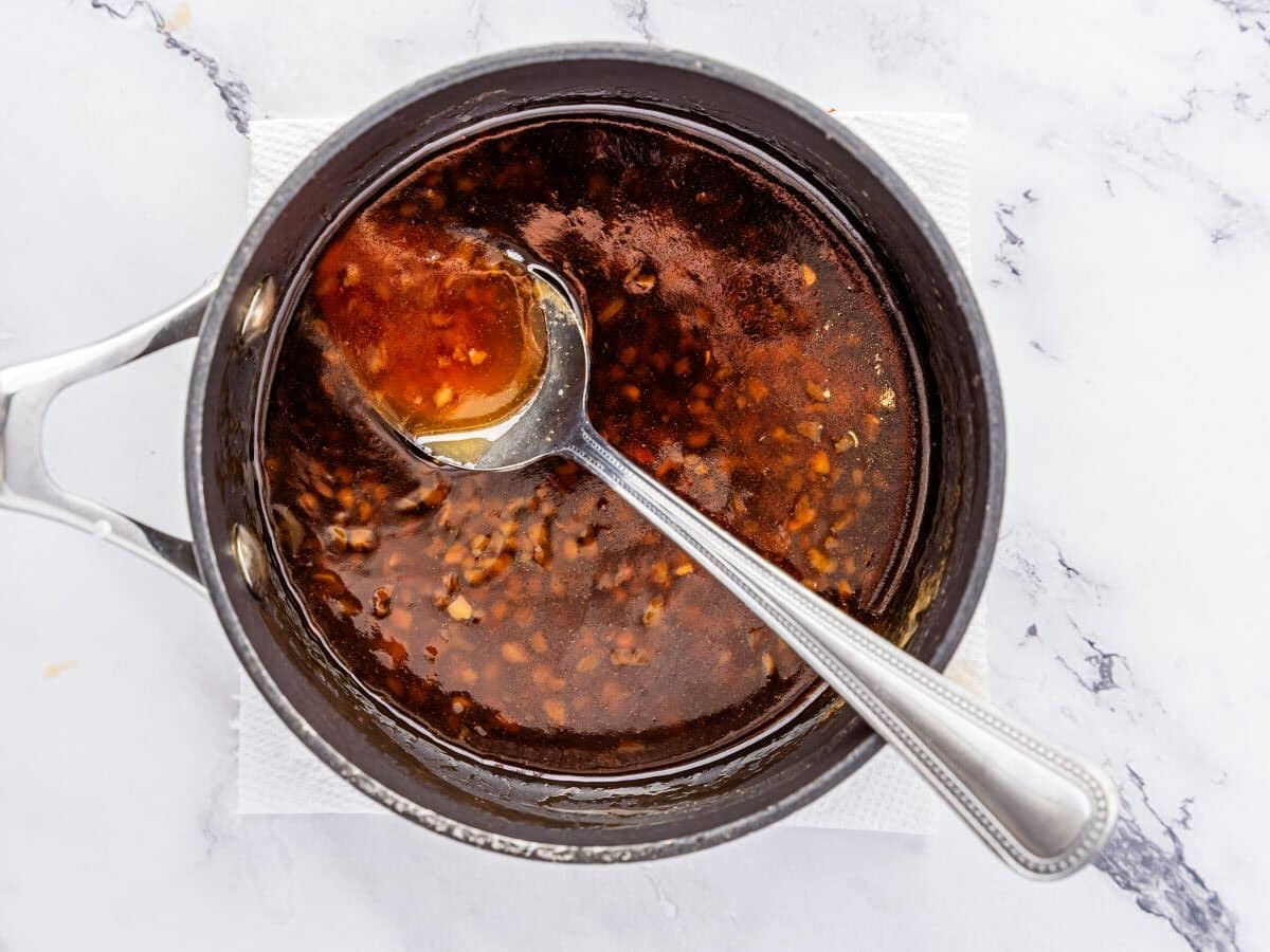 A saucepan filled with dark, glossy honey garlic sauce with visible minced garlic or ginger. A metal spoon rests in the sauce, perfect for drizzling over chicken bites, all set on a white paper towel atop a marble surface.