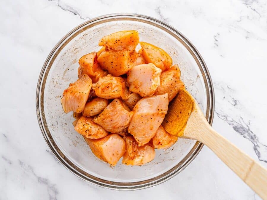 A glass bowl filled with seasoned, uncooked chicken bites is being stirred with a wooden spoon on a white marble surface, ready to be coated in honey garlic sauce.