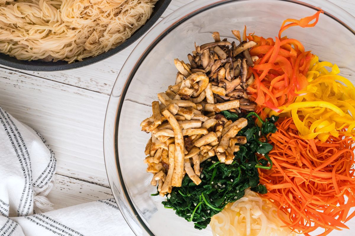 A glass bowl holds neatly arranged piles of sautéed mushrooms, tofu strips, spinach, julienned carrots, and yellow bell peppers—perfect for Tofu Japchae with Vermicelli. Next to it is a pan of cooked noodles on a white wooden surface with a striped kitchen towel.