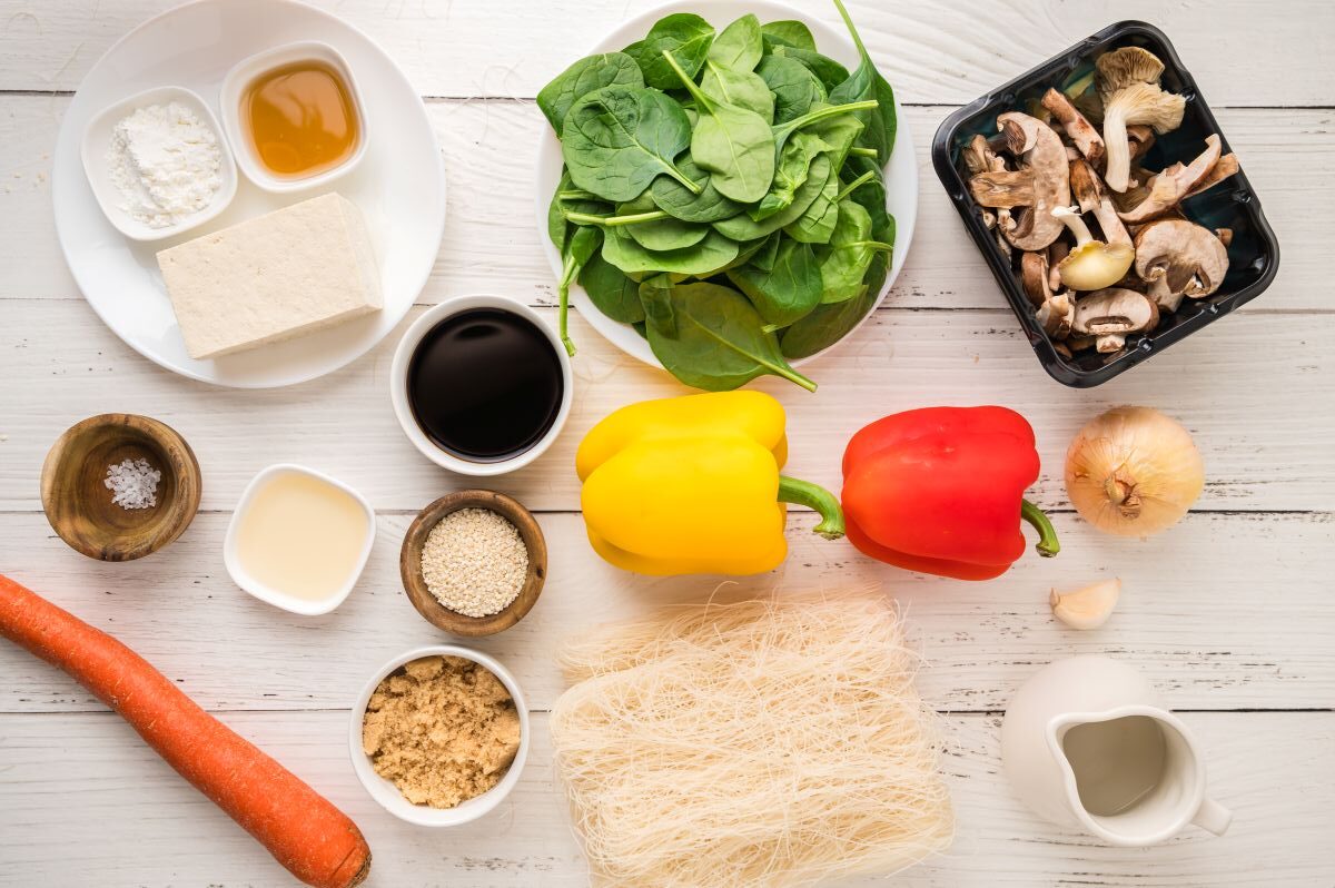 A flat lay of ingredients for Tofu Japchae with Vermicelli on a white table, featuring tofu, honey, soy sauce, vinegar, brown sugar, sesame seeds, carrot, spinach, mushrooms, bell peppers, onion, garlic, glass noodles, and a jug of liquid.