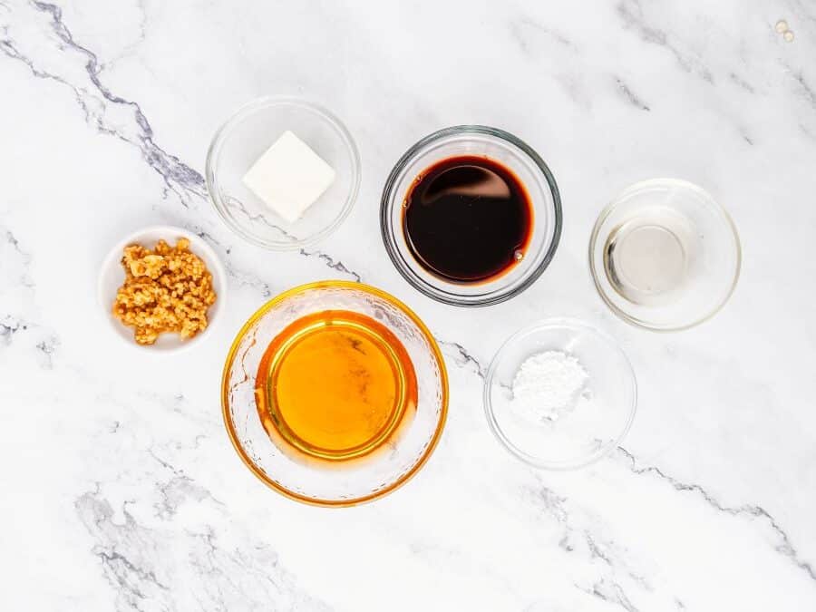 Six small glass bowls on a white marble surface, each holding ingredients—minced garlic, butter, soy sauce, rice vinegar, honey, and cornstarch—perfect for making chicken bites with a flavorful honey garlic sauce.