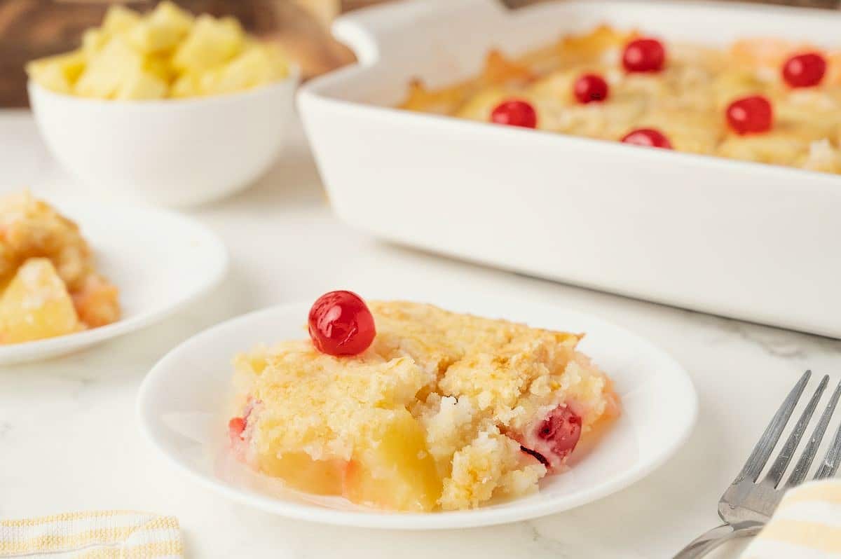 A slice of pineapple cherry dump cake sits on a white plate, topped with a red cherry. In the background, there is a baking dish with more cake and a bowl of pineapple chunks. A fork rests nearby on the table.