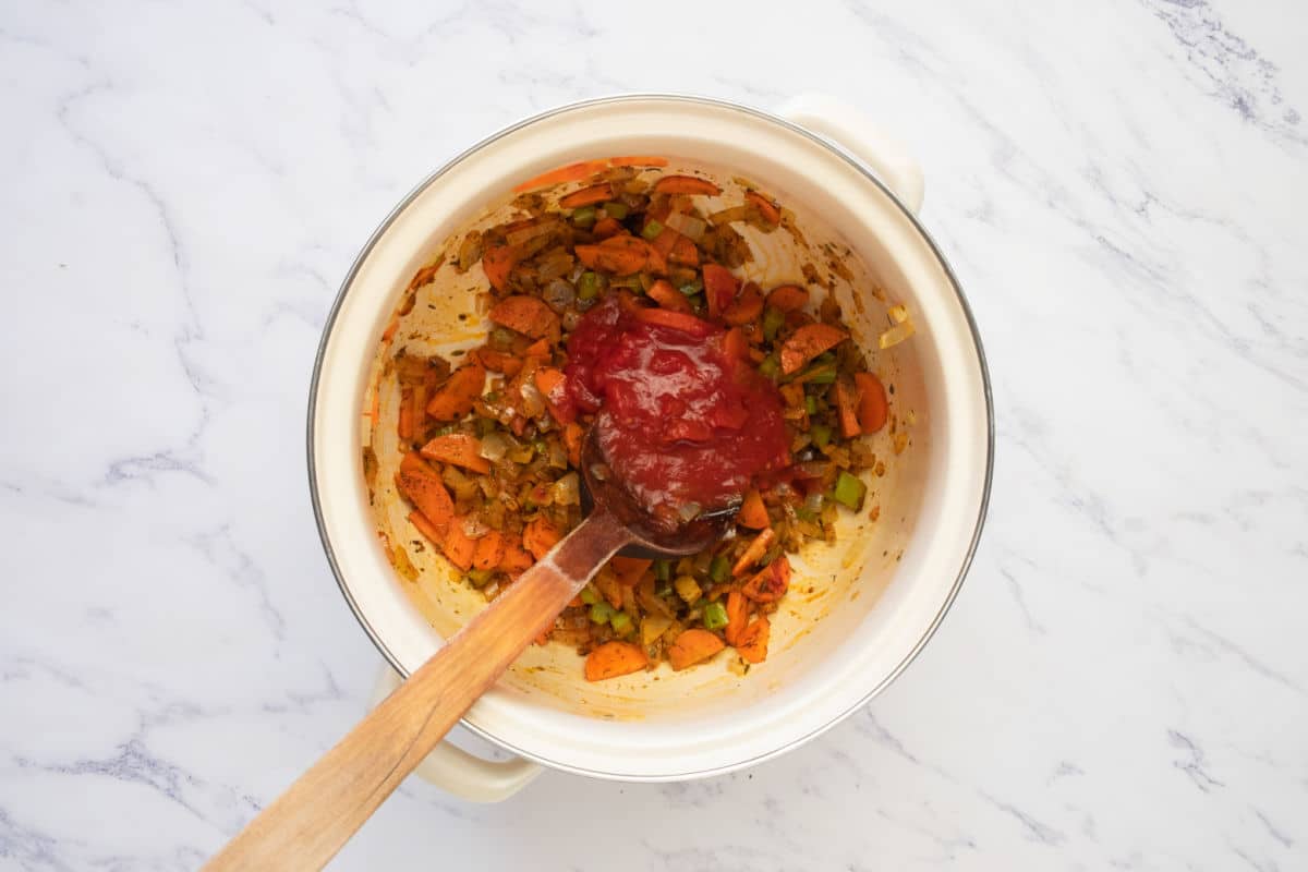 A white pot contains chopped carrots, celery, onions, and a large dollop of tomato paste, with a wooden spoon resting inside, all on a marble surface.