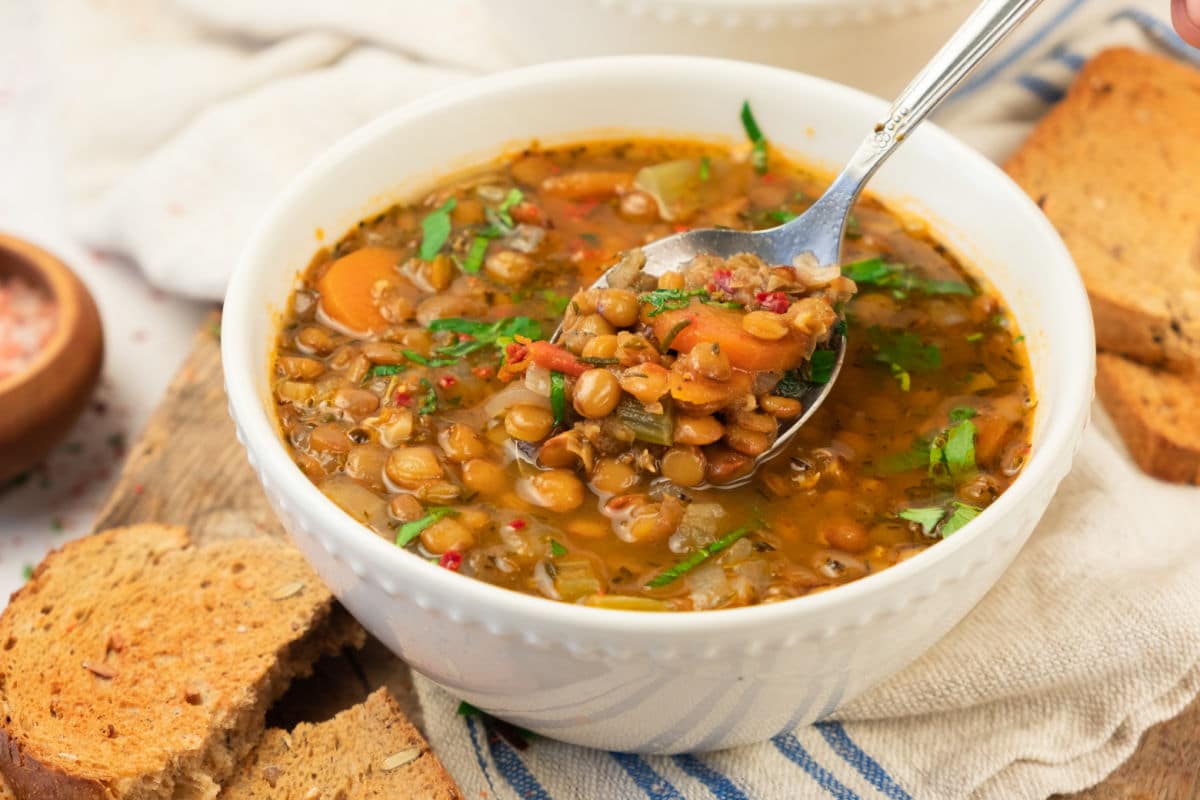 A white bowl filled with hearty lentil soup with vegetables, garnished with herbs. A spoon is lifting some soup from the bowl, and slices of toasted bread are nearby on a wooden board.
