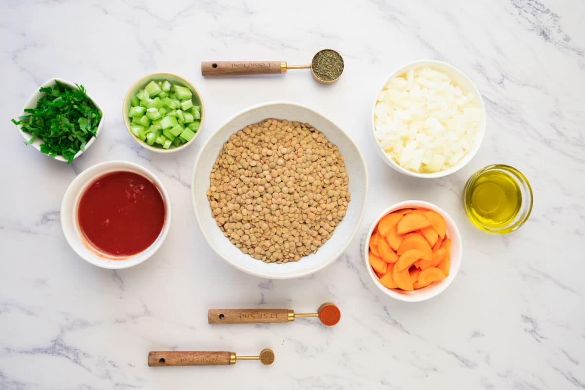 A top-down view of bowls containing uncooked lentils, chopped onions, sliced carrots, celery, parsley, tomato sauce, olive oil, and measuring spoons with various spices on a white marble surface.