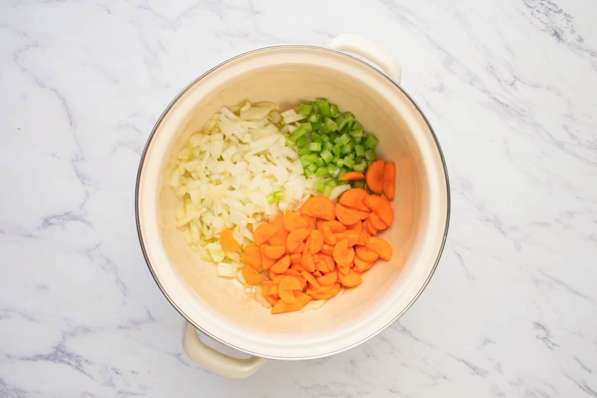 A cream-colored pot on a marble surface contains chopped onions, celery, and carrots arranged in separate sections.