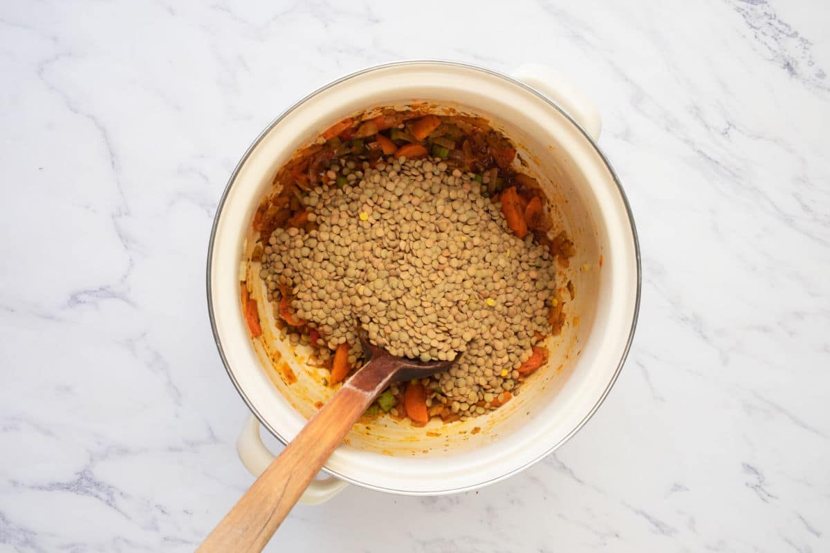 A white pot on a marble surface contains uncooked green lentils mixed with chopped vegetables, including carrots and celery, with a wooden spoon resting inside.