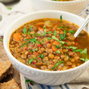 A white bowl filled with hearty lentil soup, featuring visible chopped carrots, onions, and fresh herbs, sits on a striped cloth with pieces of whole grain bread nearby. A spoon rests inside the bowl.
