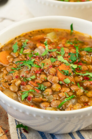 A white bowl filled with hearty lentil soup, featuring visible chopped carrots, onions, and fresh herbs, sits on a striped cloth with pieces of whole grain bread nearby. A spoon rests inside the bowl.