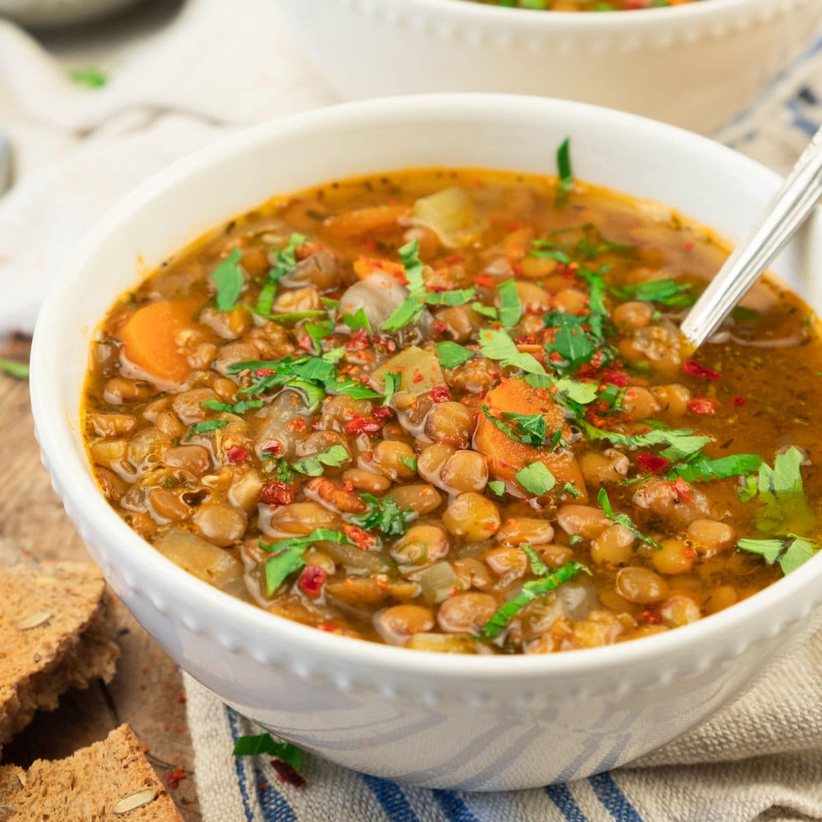 A white bowl filled with hearty lentil soup, featuring visible chopped carrots, onions, and fresh herbs, sits on a striped cloth with pieces of whole grain bread nearby. A spoon rests inside the bowl.