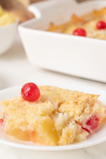 A slice of pineapple cherry cobbler sits on a white plate, topped with a red cherry. More cobbler is in a white baking dish in the background, with a bowl of pineapple chunks and a fork nearby.