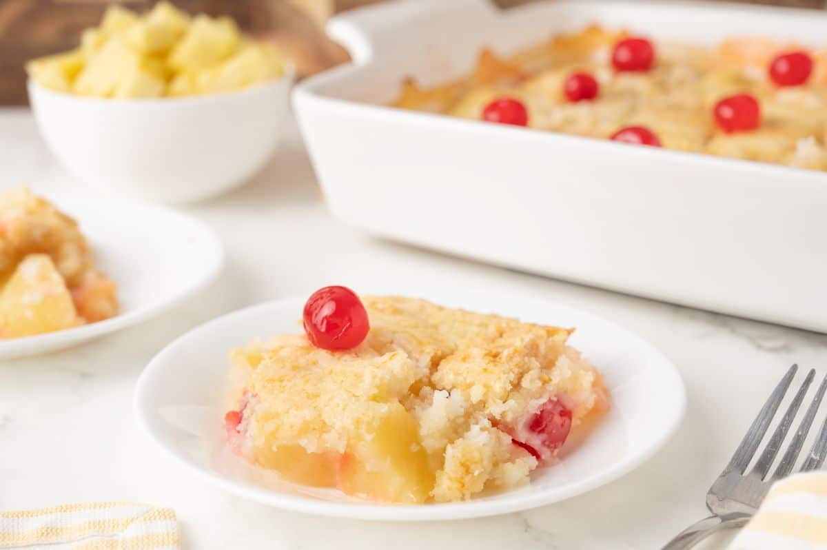 A slice of pineapple cherry cobbler sits on a white plate, topped with a red cherry. More cobbler is in a white baking dish in the background, with a bowl of pineapple chunks and a fork nearby.