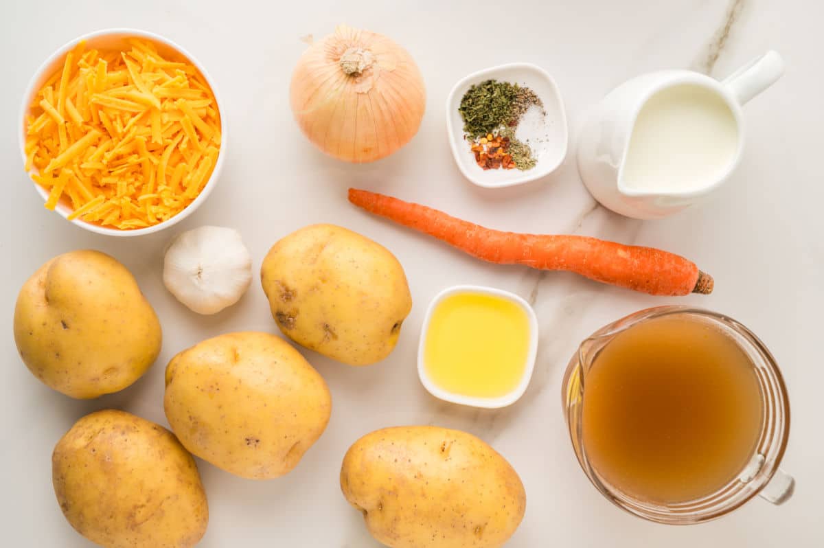 A flat lay of ingredients for potato soup, including potatoes, shredded cheddar cheese, an onion, a carrot, a garlic bulb, olive oil, vegetable broth, milk, and small bowls of spices on a white surface.