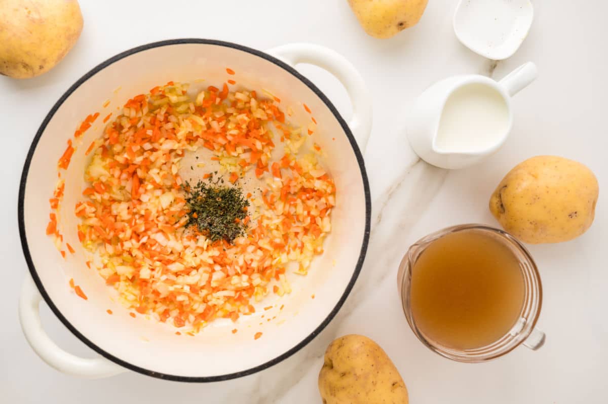 Top view of a pot with diced carrots, onions, and herbs simmering for potato soup; surrounded by whole potatoes, a glass measuring cup of broth, and a small jug of cream on a white surface.