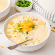 A bowl of creamy potato soup topped with shredded cheddar cheese and chopped green onions, with a spoon resting inside. A small bowl of extra green onions and a striped towel sit in the background.