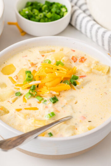 A bowl of creamy potato soup topped with shredded cheddar cheese and chopped green onions, with a spoon resting inside. A small bowl of extra green onions and a striped towel sit in the background.