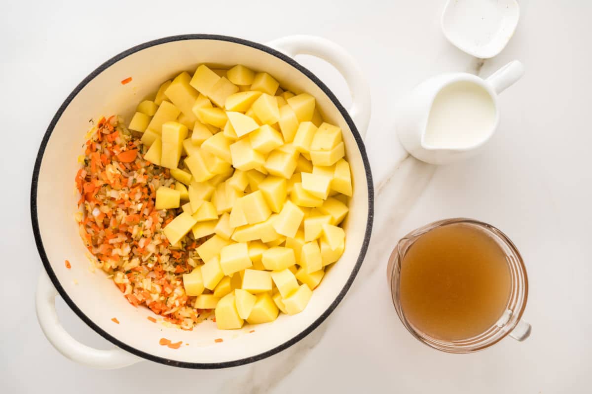 A white pot filled with diced potatoes and sautéed vegetables, ready for creamy potato soup, sits on a white surface. Next to it are a glass pitcher of broth and a small white pitcher of cream.
