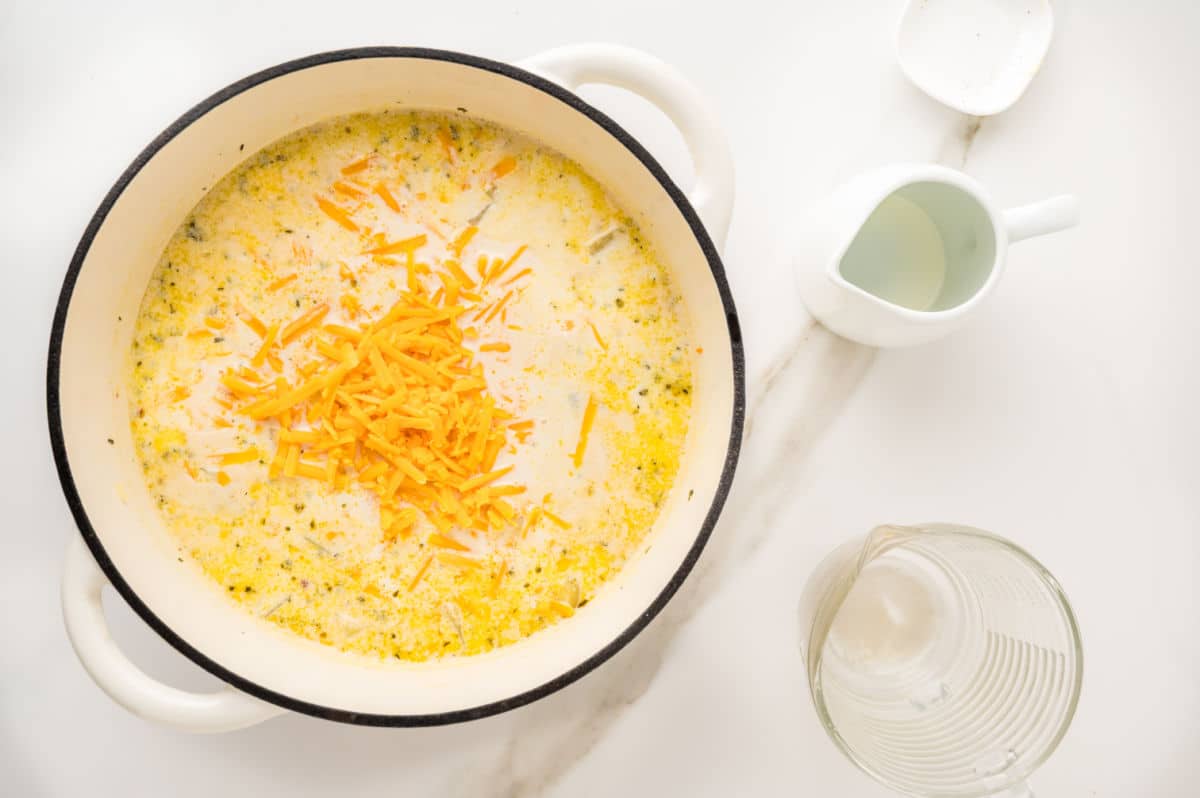A white pot filled with creamy potato soup topped with shredded cheddar cheese sits on a white surface, surrounded by a clear measuring cup and a small white pitcher.