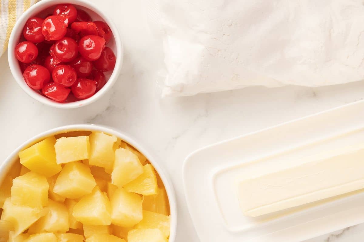 A bowl of maraschino cherries, a bowl of pineapple chunks, a stick of butter on a dish, and a bag of flour on a white countertop.
