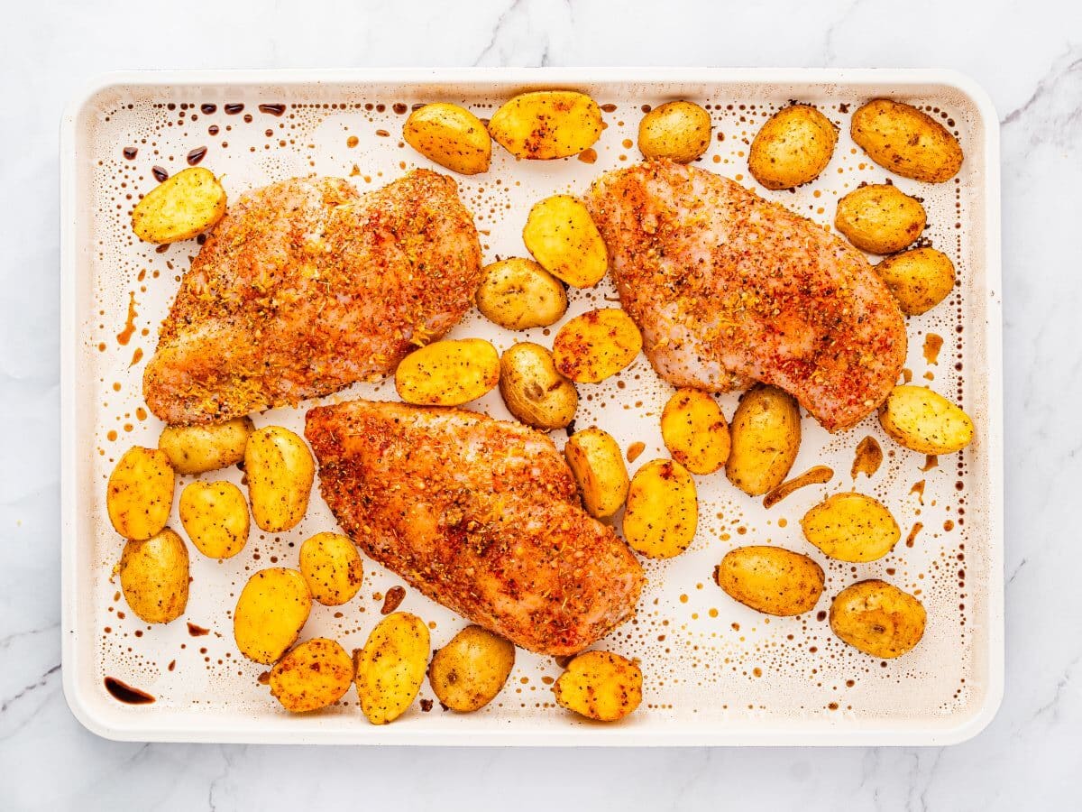 A baking sheet with three seasoned chicken breasts and halved roasted baby potatoes on a white marble surface. The food is evenly spaced and sprinkled with spices.