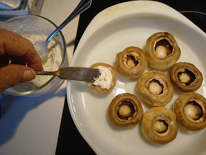 A hand spreads creamy filling into a raw, hollowed-out mushroom cap on a white plate with several other empty mushroom caps nearby.