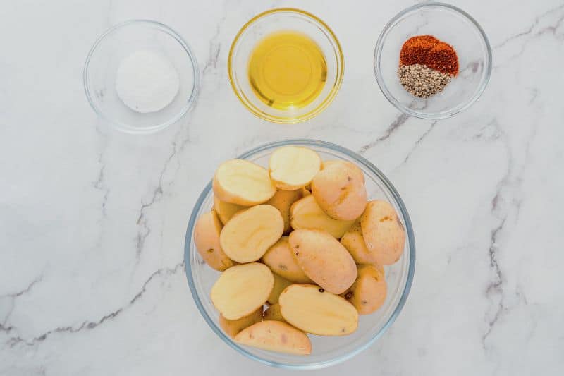 A bowl of halved potatoes is surrounded by small bowls containing oil, salt, and a mix of black pepper with red spices, all placed on a white marble surface.