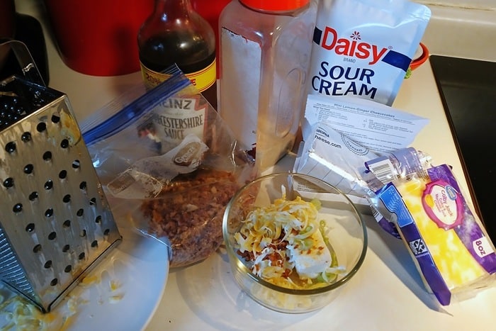 A bowl with shredded cheese, bacon bits, and sour cream sits on a counter surrounded by a cheese grater, sour cream, Worcestershire sauce, bacon bits, and shredded cheese packaging.