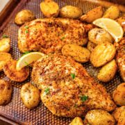 Seasoned baked chicken breasts and roasted baby potatoes on a textured baking sheet, garnished with lemon wedges and sprinkled herbs.