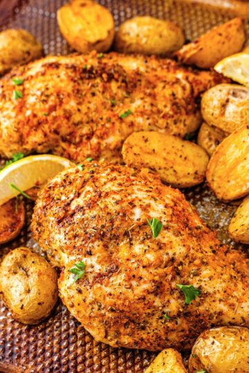 Seasoned baked chicken breasts and roasted baby potatoes on a textured baking sheet, garnished with lemon wedges and sprinkled herbs.