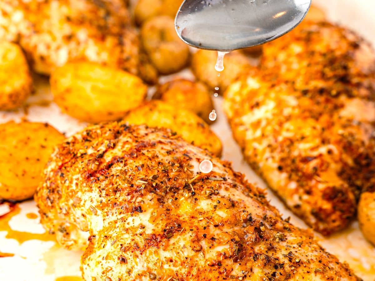 Seasoned Sheet-Pan Chicken breasts and crispy potatoes on a tray, with a spoon drizzling sauce or juices over the chicken, highlighting the texture and spices.