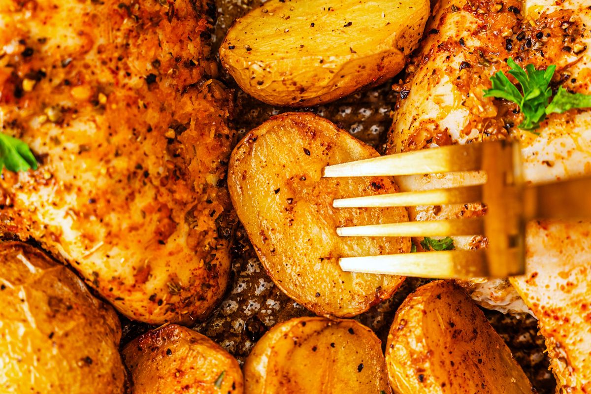 Close-up of golden roasted potatoes and seasoned chicken, with a fork piercing one potato slice. The food is garnished with herbs and cooked to a crispy, appetizing finish.