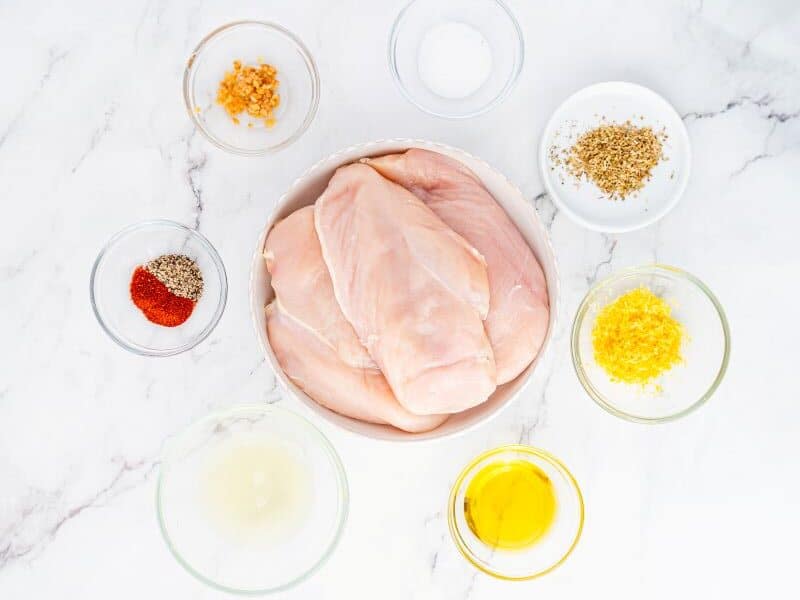 Raw chicken breasts in a bowl surrounded by small bowls of minced garlic, salt, dried herbs, lemon zest, black pepper with paprika, olive oil, and lemon juice on a white marble surface.